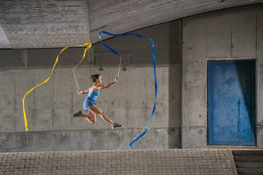 Junge Frau hält Bänder und übt rhythmische Gymnastik an einer Betonwand unter einer Brücke - STSF02565