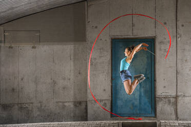 Young woman holding ribbon jumping while doing aerobics against concrete wall - STSF02570