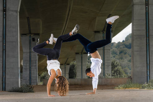 Young man and woman doing handstands on road under bridge - STSF02573