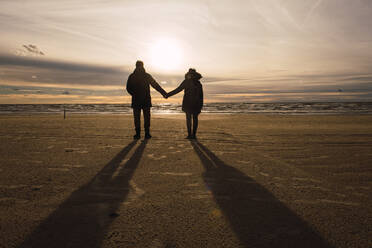 Blick auf ein Paar in Freizeitkleidung, das in der kalten Jahreszeit am Sandstrand steht und sich an den Händen hält, beleuchtet - ADSF01607