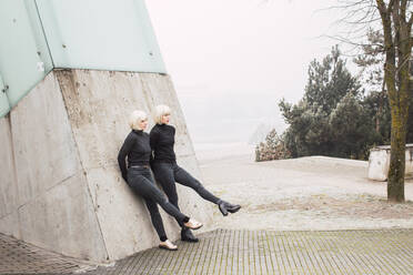 Attraktive junge Frauen in dunkler Kleidung, die ihre Beine ausstrecken und sich an eine Mauer auf einer Straße in der Nähe von Bäumen in Litauen lehnen - ADSF01609