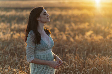 Young woman with eyes closed standing amidst wheat crops in farm at sunset - JMPF00212