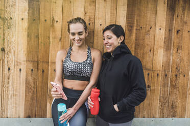 Smiling woman showing smart phone to female friend against wooden wall - DSIF00043