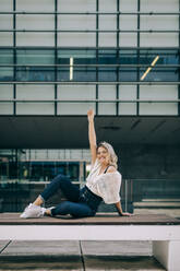 Woman with hand raised sitting on bench against office building - DCRF00423