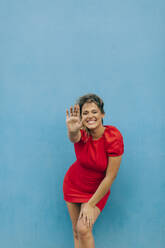 Smiling woman showing stop gesture while standing against blue wall - DCRF00445