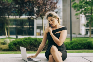 Geschäftsfrau, die mit einem Smartphone spricht, während sie einen Laptop im Freien benutzt - DCRF00454