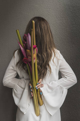 Woman with brown hair holding bouquet behind back by wall stock photo