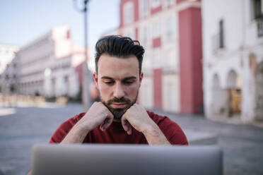 Thoughtful man sitting with hands on chin looking at laptop in city - GRCF00278