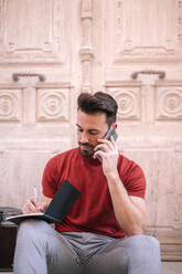 Geschäftsmann, der im Freien sitzend mit seinem Handy telefoniert und Notizen macht - GRCF00293