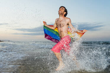 Lachender Mann, der im spritzenden Wasser rennt und eine Flagge des Schwulenstolzes trägt - CJMF00314