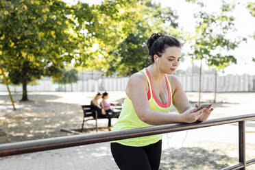 Junge Frau in Sportkleidung benutzt ihr Smartphone, während sie sich im Park an ein Geländer lehnt - CAVF87259