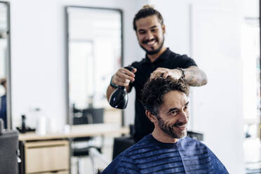 Fröhlicher männlicher Friseur, der das Haar eines lächelnden Kunden anfeuchtet - CAVF87299