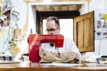 Male artist sewing fabric at table in workshop - CAVF87315