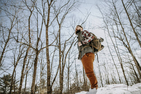 Mann mit Flanell, Bart und Rucksack steht im Winter im Schnee im Wald - CAVF87330