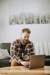 Man with beard holds cup of coffee while working on laptop computer - CAVF87337