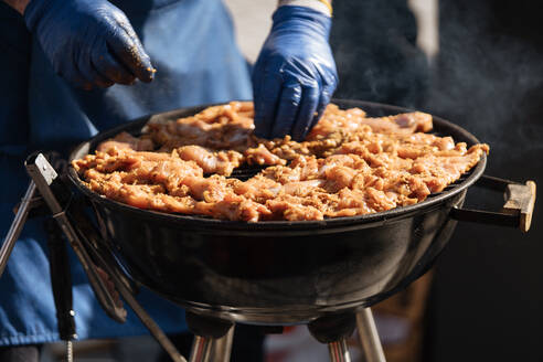 Gesichtslose Aufnahme eines Mannes mit Handschuhen, der rohes Fleisch auf einem Metallgitter in einem im Sonnenlicht stehenden Grill anrichtet - ADSF01661