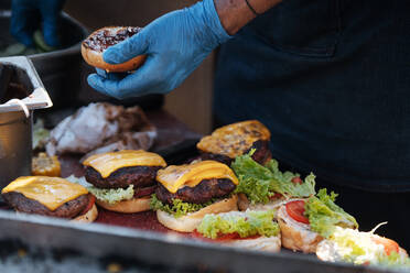 Gesichtslose Aufnahme eines Kochs mit Handschuhen, der Brötchen mit frischem Gemüse und Fleischpasteten mit Käse anrichtet und Burger zubereitet - ADSF01664