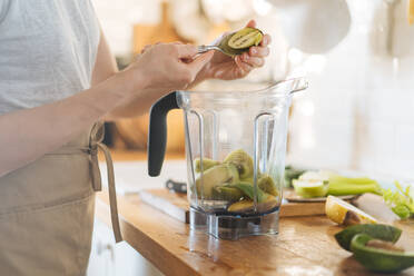 Crop side view of woman peeling kiwi half with spoon and mixing with other fruit in blender bowl for green smoothie - ADSF01670