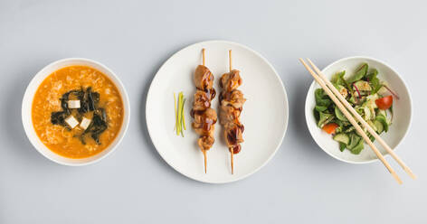 Beautiful set of bowls with miso soup and fresh vegetable salad and plate with sushi rolls and wooden chopsticks lying on white background from above - ADSF01672