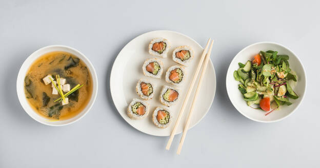 Schönes Set von Schalen mit Misosuppe und frischem Gemüsesalat und Teller mit Sushi-Rollen und hölzernen Essstäbchen auf weißem Hintergrund von oben liegend - ADSF01673