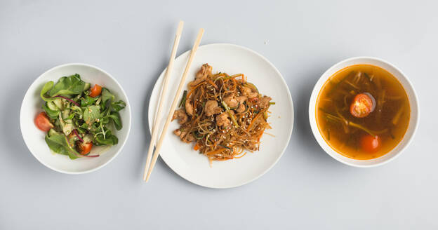 Schönes Set von Schalen mit Misosuppe und frischem Gemüsesalat und Teller mit Sushi-Rollen und hölzernen Essstäbchen auf weißem Hintergrund von oben liegend - ADSF01674