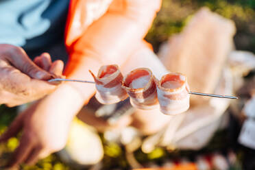 Faceless shot of person arranging folded bacon strips on metal skewer for barbecue meal - ADSF01688