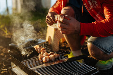 Mann bereitet Speck und Würstchen auf Spießen vor, die auf brennender Holzkohle in einem tragbaren Grill im Freien gegrillt werden - ADSF01689