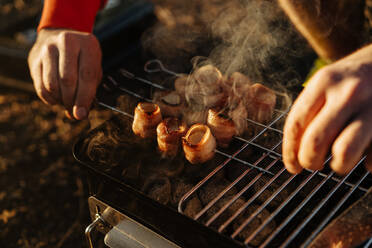 Man preparing bacon and sausages on skewers grilling on burning charcoal in portable griddle outside - ADSF01690