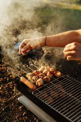 Crop woman preparing bacon and sausages on skewers grilling on burning charcoal in portable griddle outside - ADSF01692