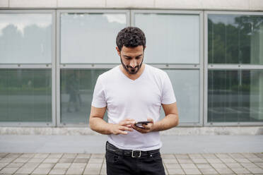 Handsome mid adult man using smart phone while standing on footpath against building - MEUF01517