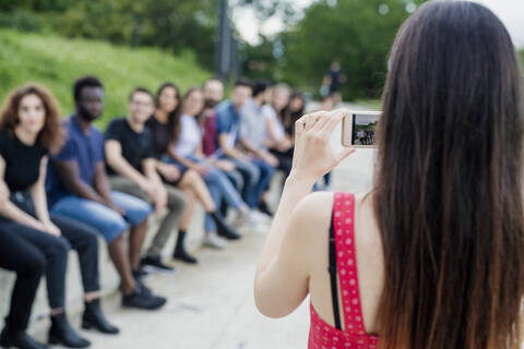 Woman photographing friends sitting in row with smart phone at park stock photo