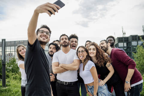 Cheerful man taking selfie with group of friends in park against sky stock photo