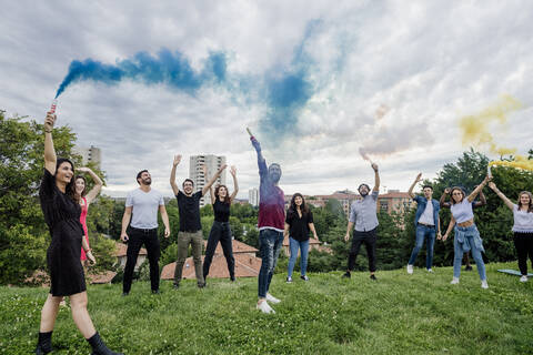 Cheerful group of friends playing with smoke bombs in park against cloudy sky stock photo