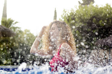 Side view of curly redhead woman adjusting hair and standing in pool with splashes - ADSF01743