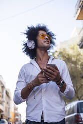 Attractive and latino man with afro hair and yellow pilot glasses on a street and listen music with headphones and smart phone and smile - ADSF01752
