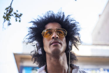 Attraktiver und Latino-Mann mit Afro-Haar und gelber Pilotenbrille auf einer Straße und hören Musik mit Kopfhörern und Smart Phone - ADSF01753