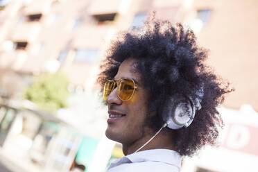 Attraktiver und Latino-Mann mit Afro-Haar und gelber Pilotenbrille auf einer Straße und hören Musik mit Kopfhörern und Smartphone und Lächeln - ADSF01754