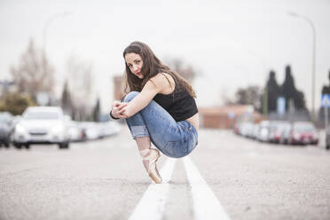 Woman dancer on ballet tips and jeans on the street and hugging his knees - ADSF01776