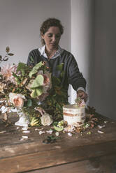 Seitenansicht der Dame Platzierung Gericht mit leckeren Kuchen dekoriert Blüte Knospe auf Holztisch mit Strauß Chrysanthemen, Rosen und Pflanze Zweige in Vase zwischen trockenen Blättern auf grauem Hintergrund - ADSF01819