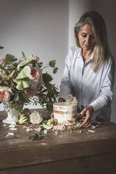 Seitenansicht der Dame Platzierung Gericht mit leckeren Kuchen dekoriert Blüte Knospe auf Holztisch mit Strauß Chrysanthemen, Rosen und Pflanze Zweige in Vase zwischen trockenen Blättern auf grauem Hintergrund - ADSF01828