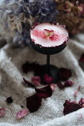 Beautiful Nordic drink in small high stem glass sprinkled with red powder and leaf served on grey cloth with scattered dried rose petals on blurred background - ADSF01866