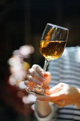 Schnittansicht einer Frau in gestreifter Bluse, die ein elegantes Glas Weißwein hält, das an einem Holztisch mit glitzernden Getränken auf einem unscharfen Hintergrund steht - ADSF01932