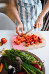 Crop-Ansicht einer Frau in Sommerkleidung, die leckere frische Tomaten auf einem Holzbrett schneidet, während andere Zutaten auf dem Tisch liegen - ADSF01944