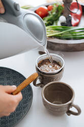 Crop hands pouring hot water from kettle into beautiful ceramic mug with small filter scoop in it standing on table with cooking ingredients - ADSF01946