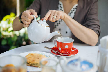 Schnittansicht einer Frau, die eine Porzellankanne in der Hand hält und heißen Tee in eine rote, gepunktete Keramiktasse mit Untertasse und Löffel gießt, die auf einem weißen Tisch mit einem Stück Kuchen auf einem Teller in der Nähe steht - ADSF01955