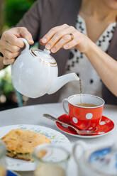 Schnittansicht einer Frau, die eine Porzellankanne in der Hand hält und heißen Tee in eine rote, gepunktete Keramiktasse mit Untertasse und Löffel gießt, die auf einem weißen Tisch mit einem Stück Kuchen auf einem Teller in der Nähe steht - ADSF01956