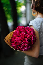 Rückenansicht einer Frau in lässiger Sommerkleidung mit Vintage-Haarschnitt und Accessoires stehen auf der Straße Bürgersteig mit lebendigen rosa auffälligen Blumen in Papier mit Bäumen auf unscharfen Hintergrund gewickelt - ADSF01965