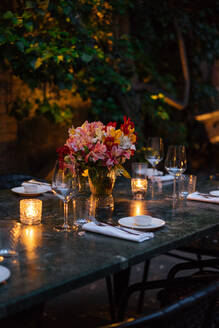 Delicate Weingläser auf dunklen Holztisch mit beleuchteten Kerzen und Blumenstrauß in der Mitte am Abend mit schönen Blumen und Bäumen um platziert - ADSF01970