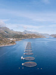 Drohnenansicht einer Reihe von Fischfarmen im blauen, ruhigen Wasser des Ozeans mit der Küstenlinie der Insel Lofoten im Sonnenlicht, Norwegen - ADSF01980