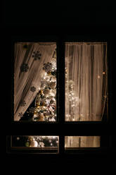 Amazing Christmas tree decorated with beautiful baubles and bright fairy lights standing behind window in cozy room at night - ADSF01997